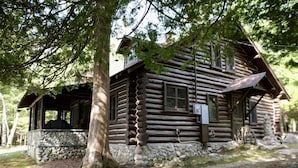 A log cabin with a stone foundation, a porch, and a large tree in front.