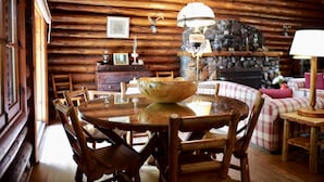 A wooden cabin interior with a round dining table, wooden chairs, a stone fireplace, and a checkered sofa.