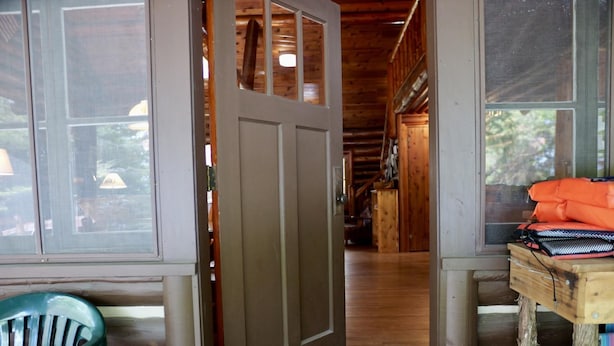 A wooden cabin interior with a door leading to a hallway, a green plastic chair, and a table with an orange bag.