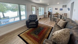 Un salon spacieux avec un canapé en cuir, un fauteuil noir et un coin repas comprenant une table et des chaises. De grandes fenêtres laissent entrer la lumière naturelle et offrent une vue sur l’extérieur.