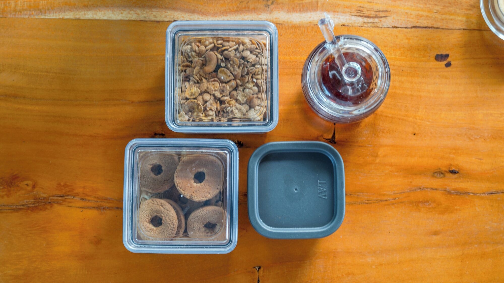 Three clear plastic containers with food items, a glass jar with a dark liquid, and a gray lid on a wooden surface.