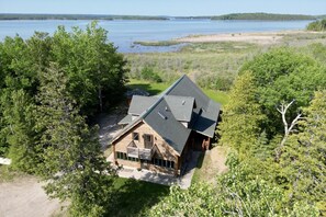 A wooden cabin with a sloped roof, surrounded by dense trees and a lake in the background.