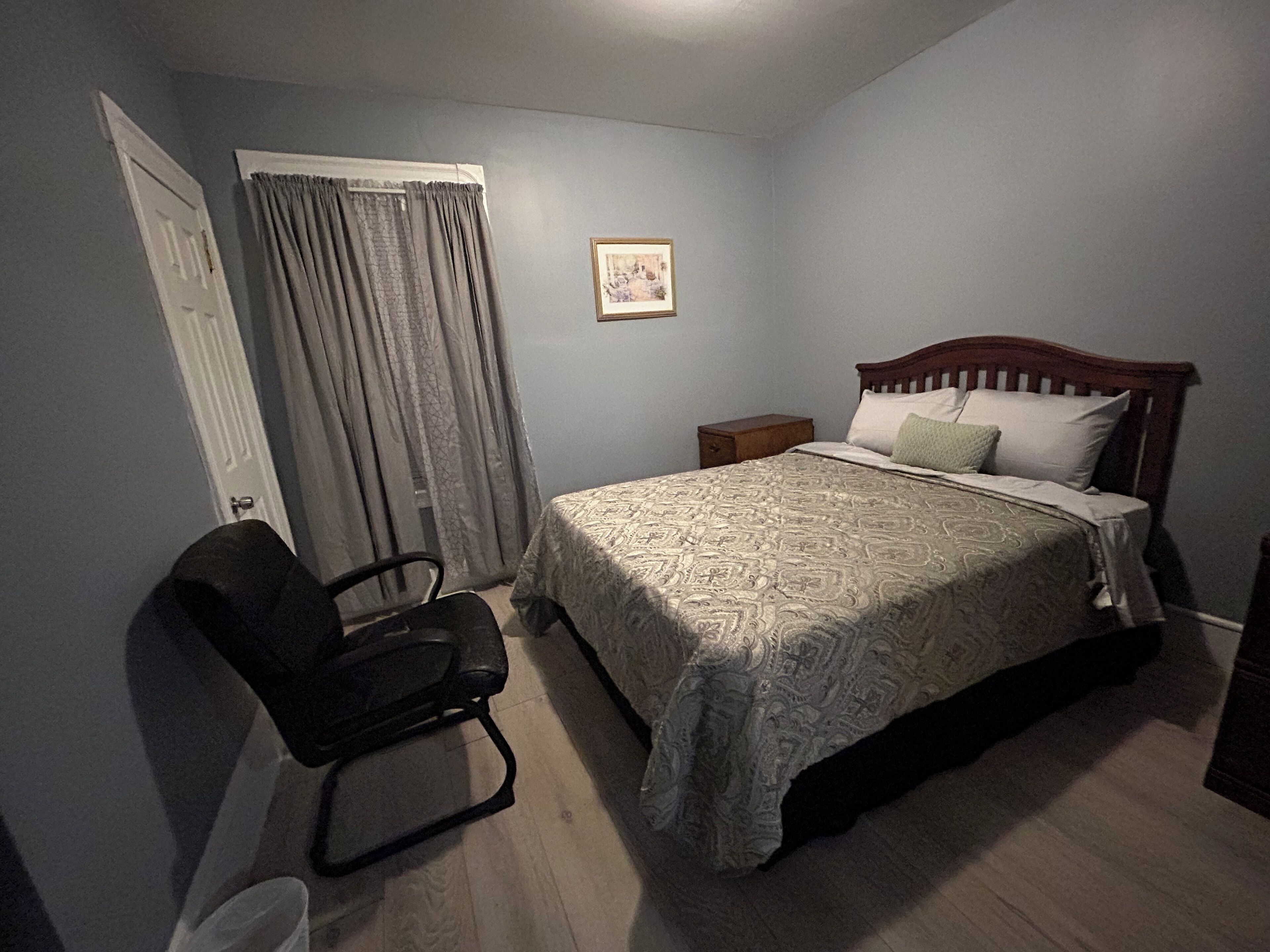 Un dormitorio con cama, una silla, una mesita de noche, un cuadro en la pared y una puerta con cortinas.