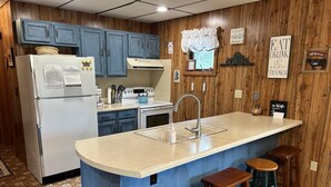A kitchen with blue cabinets, a white refrigerator, a stove, a sink, and three wooden stools.
