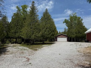 Un vialetto sterrato che conduce a un garage rosso con porta bianca, circondato da alberi alti.