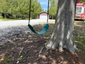 Une balançoire fixée à un arbre, dans un environnement naturel, avec un chemin de gravier et un bâtiment rouge en arrière-plan.