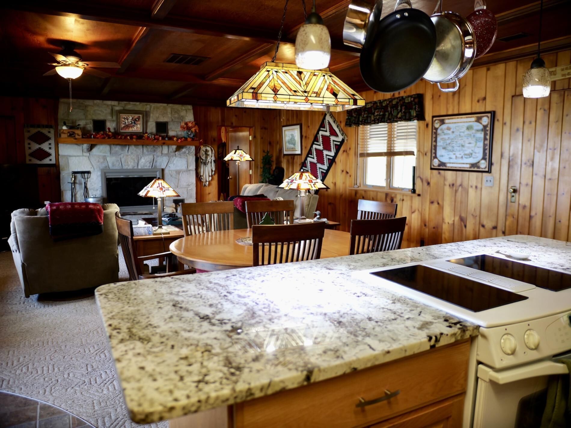 A cozy cabin-style kitchen with a granite countertop, wooden dining table, and chairs. There is a stove, a sink, and hanging pots above the counter.