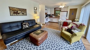 Un salon avec un canapé noir, un tapis à motifs, un pouf marron et un fauteuil jaune. Un tableau représentant des cerfs est accroché au mur.
