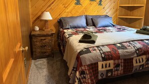 Una camera accogliente con soffitto in listelli di legno, un letto con copriletto, due cuscini, un comodino con lampada e una piccola mensola.