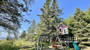 A wooden playhouse with a slide, surrounded by trees and grass.