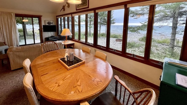 A dining area with a wooden table and chairs, a lamp, and a view of the beach through large windows.