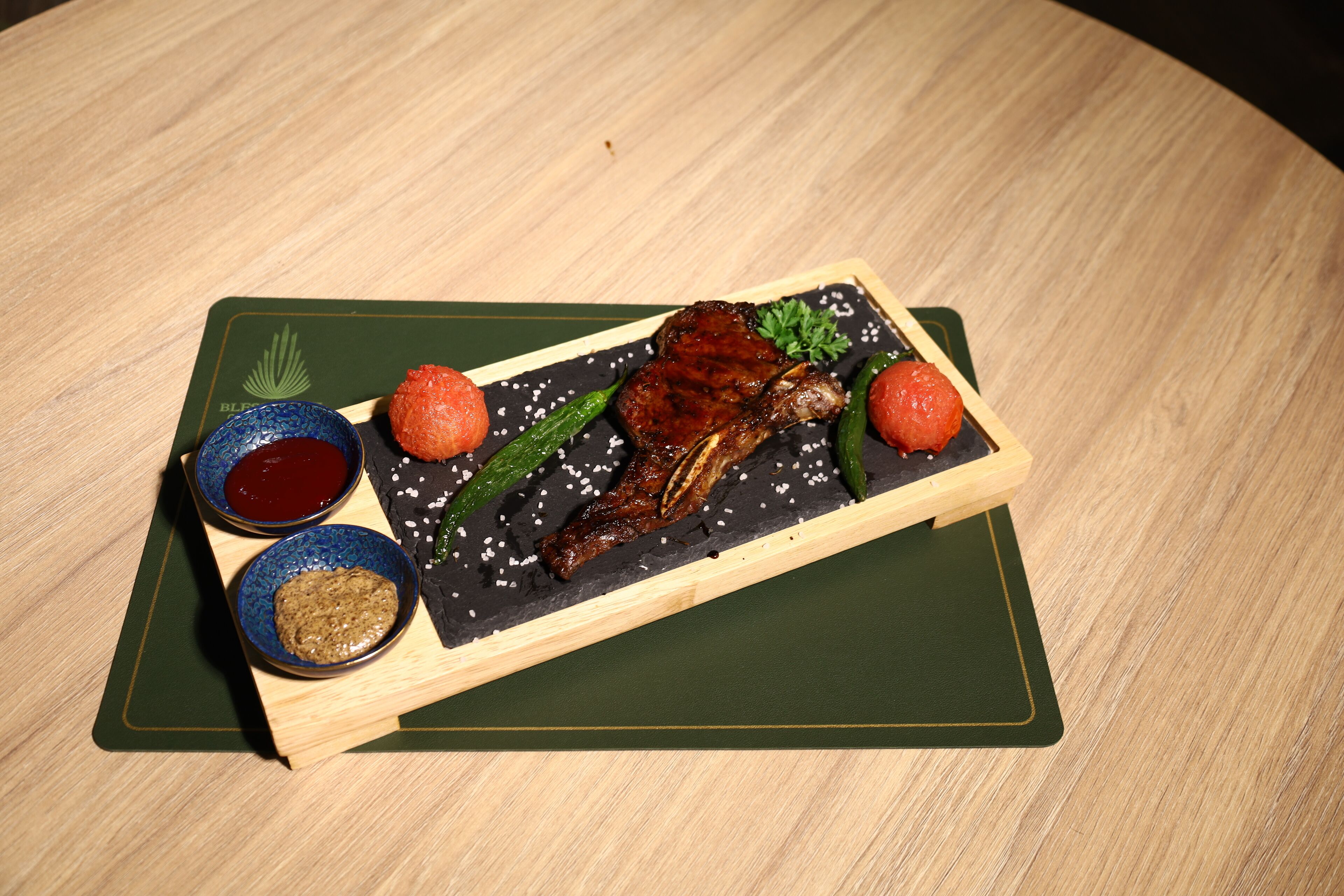 A wooden table with a dark slate serving board holding a roasted meat dish, two small bowls of sauce, and some green vegetables.