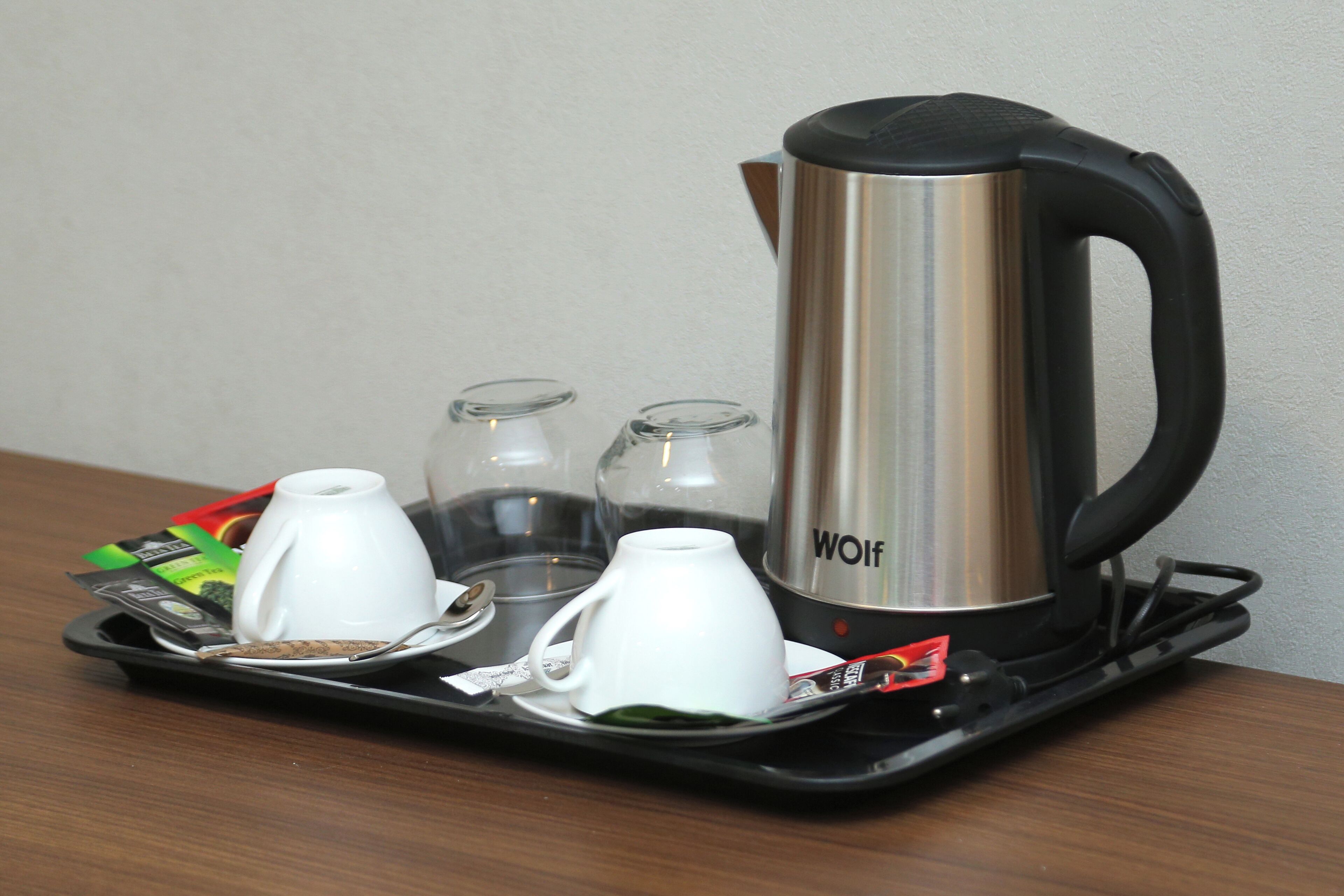 A stainless steel kettle with a black handle and the word "WOLF" on it, placed on a black tray with two white cups and two clear glasses.