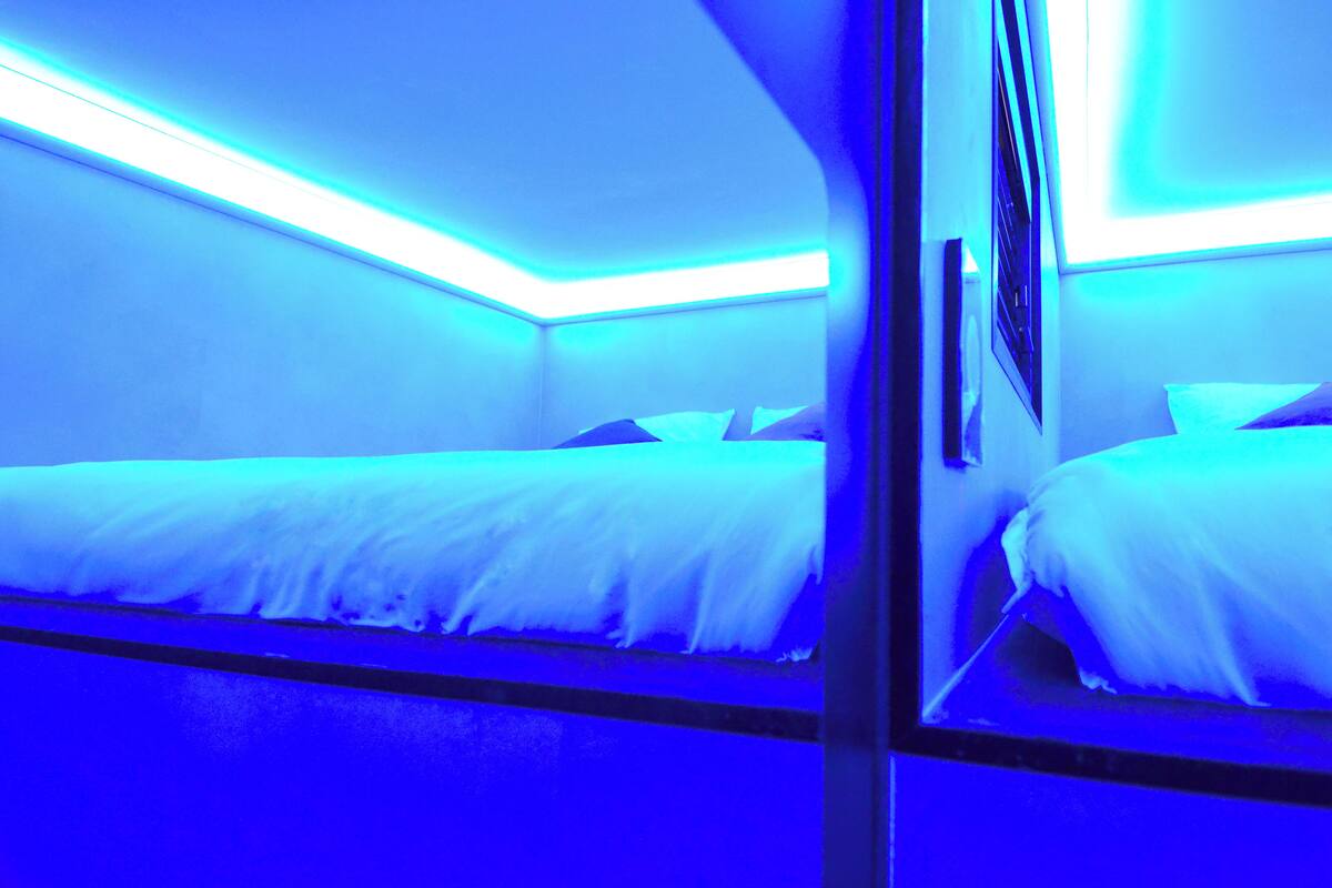 A room with two beds, each with white bedding and a headboard. The room has a modern design with blue lighting accents.