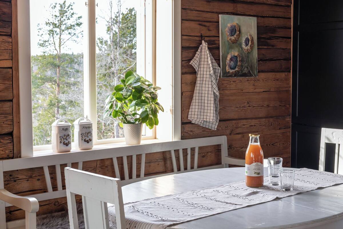 A wooden cabin interior with a table set for two, a potted plant, a painting, and a bottle of apple juice.