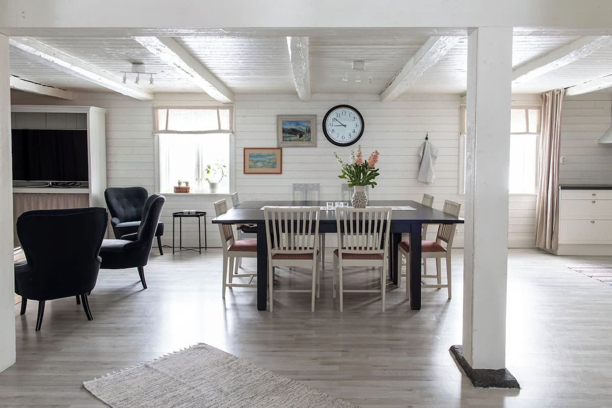 A dining area with a wooden table and chairs, a clock on the wall, and a television in the background.