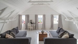 Amplia habitación en el ático, con paredes de madera blanca y techo inclinado. Incluye un sofá gris con cojines estampados, una mesa de centro de madera y un escritorio blanco con silla. Grandes ventanas con cortinas permiten la entrada de luz natural.