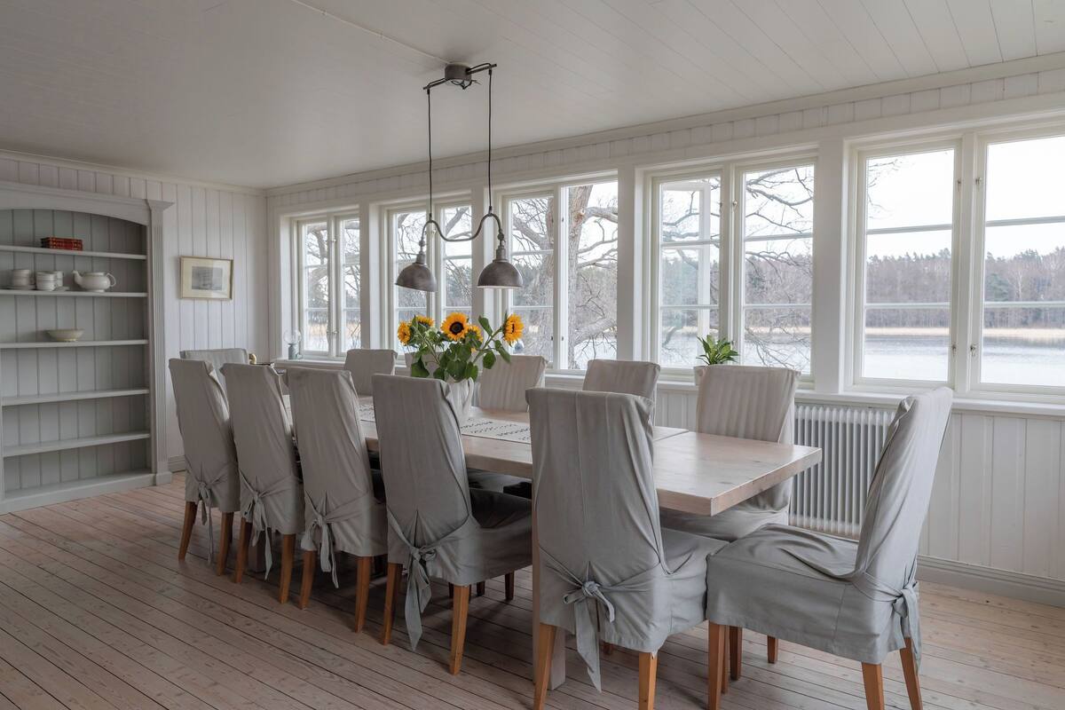 A dining room with a wooden table and chairs, large windows, and a view of a lake.