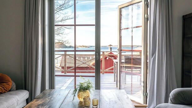 Una habitación con un gran ventanal corredizo que da a una terraza con vistas a un lago y un edificio rojo. Hay una mesa de madera con una piña y dos vasos encima.
