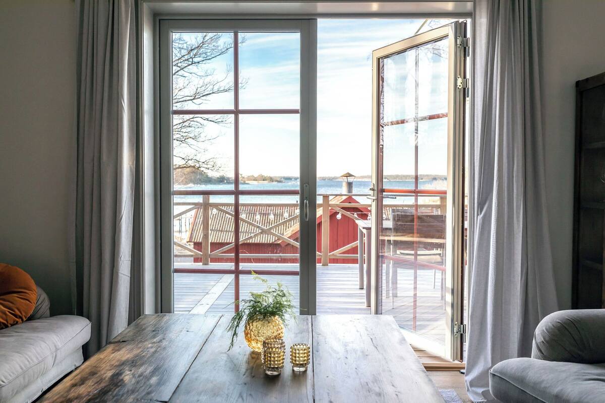 A room with a large sliding glass door leading to a deck with a view of a lake and a red building. There is a wooden table with a pineapple and two glasses on it.