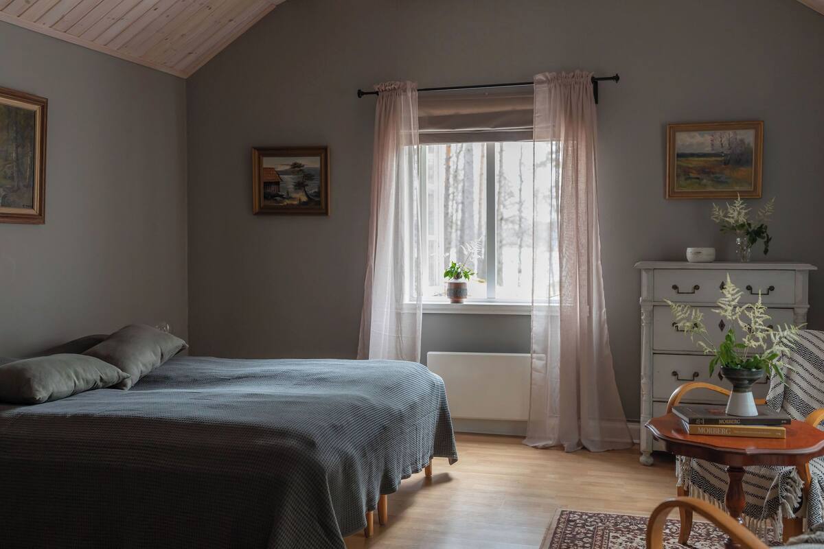 A bedroom with a bed, a dresser, a window with curtains, and a small table with a plant.