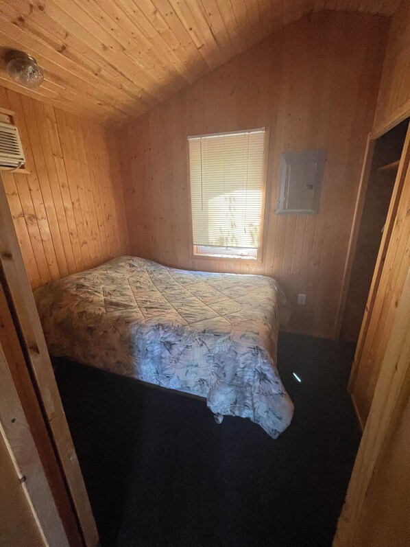 Uma pequena cabana de um único quarto, com uma cama, janela e paredes de madeira.