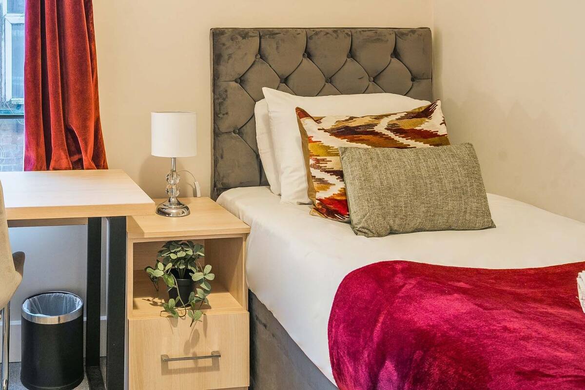 A hotel room with a bed, a desk, a chair, a radiator, a window with curtains, and a plant.