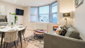 A modern living room with a grey sofa, a dining table set for four, and a large window with blinds. - Everest Lodge Serviced Apartment (Farnborough)
