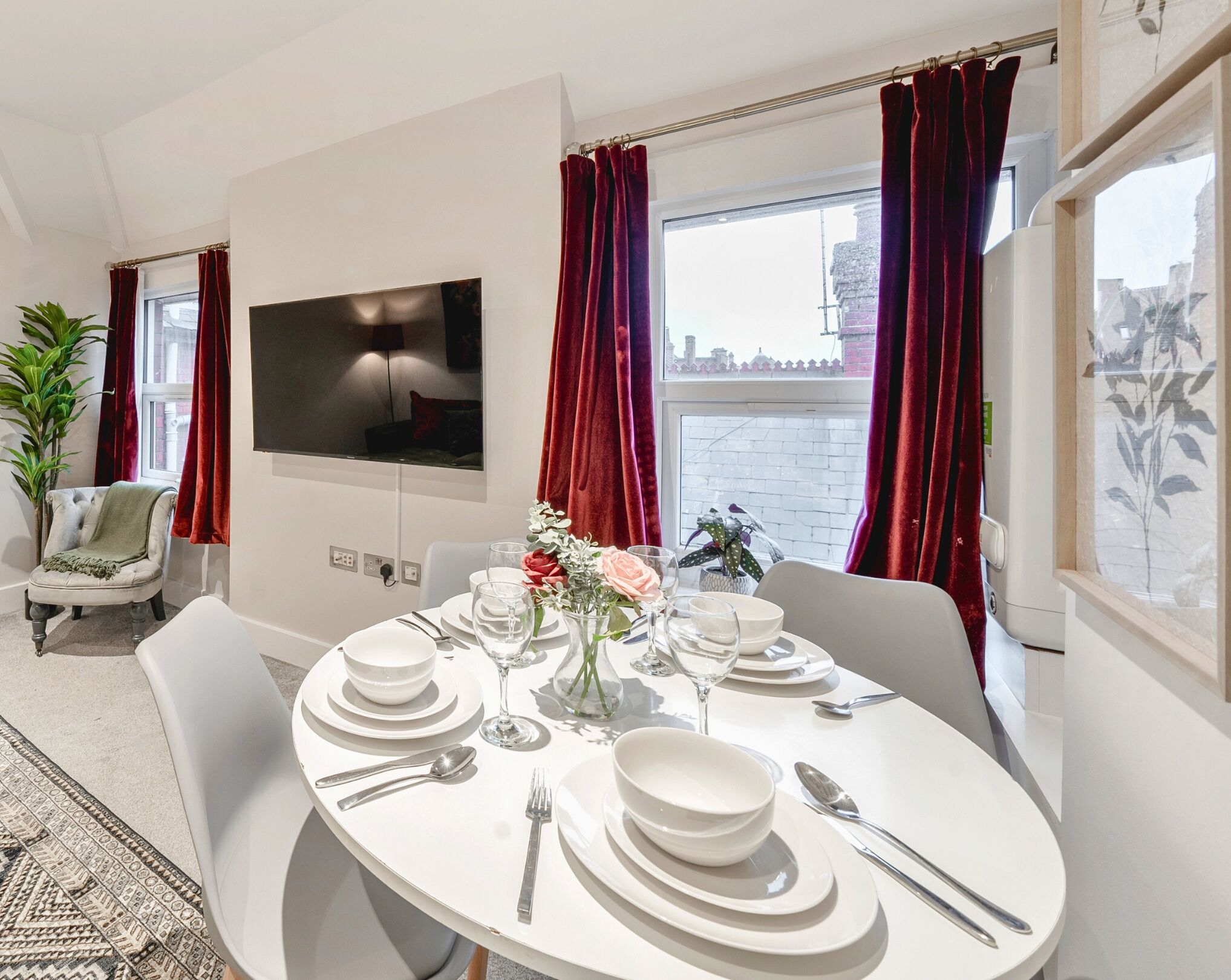 A dining area with a round table set for four, white plates, cutlery, and a vase with flowers.