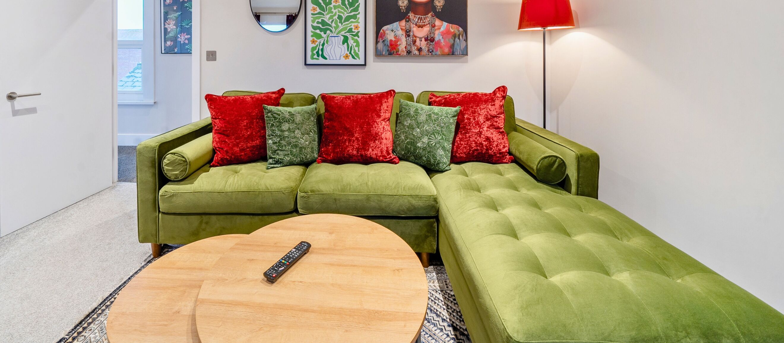 A green sofa with red and green cushions, a wooden coffee table, and a red floor lamp.