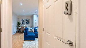 A modern living room with a blue sofa, a white door with a keypad lock, and a dining area in the background. - Everest Lodge Serviced Apartment (Farnborough)