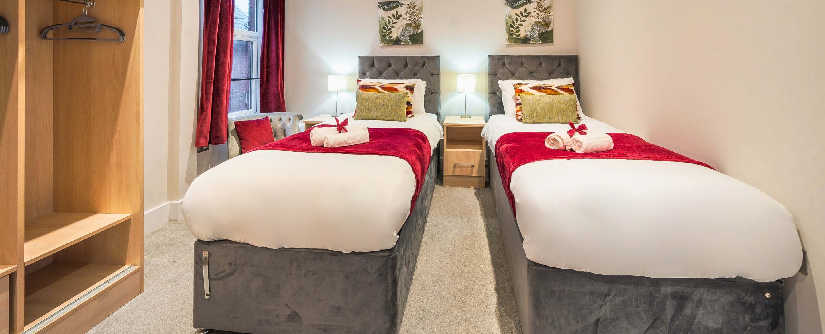 A twin room with two beds, each with a red blanket and pillows, a wooden wardrobe, and a window with red curtains.