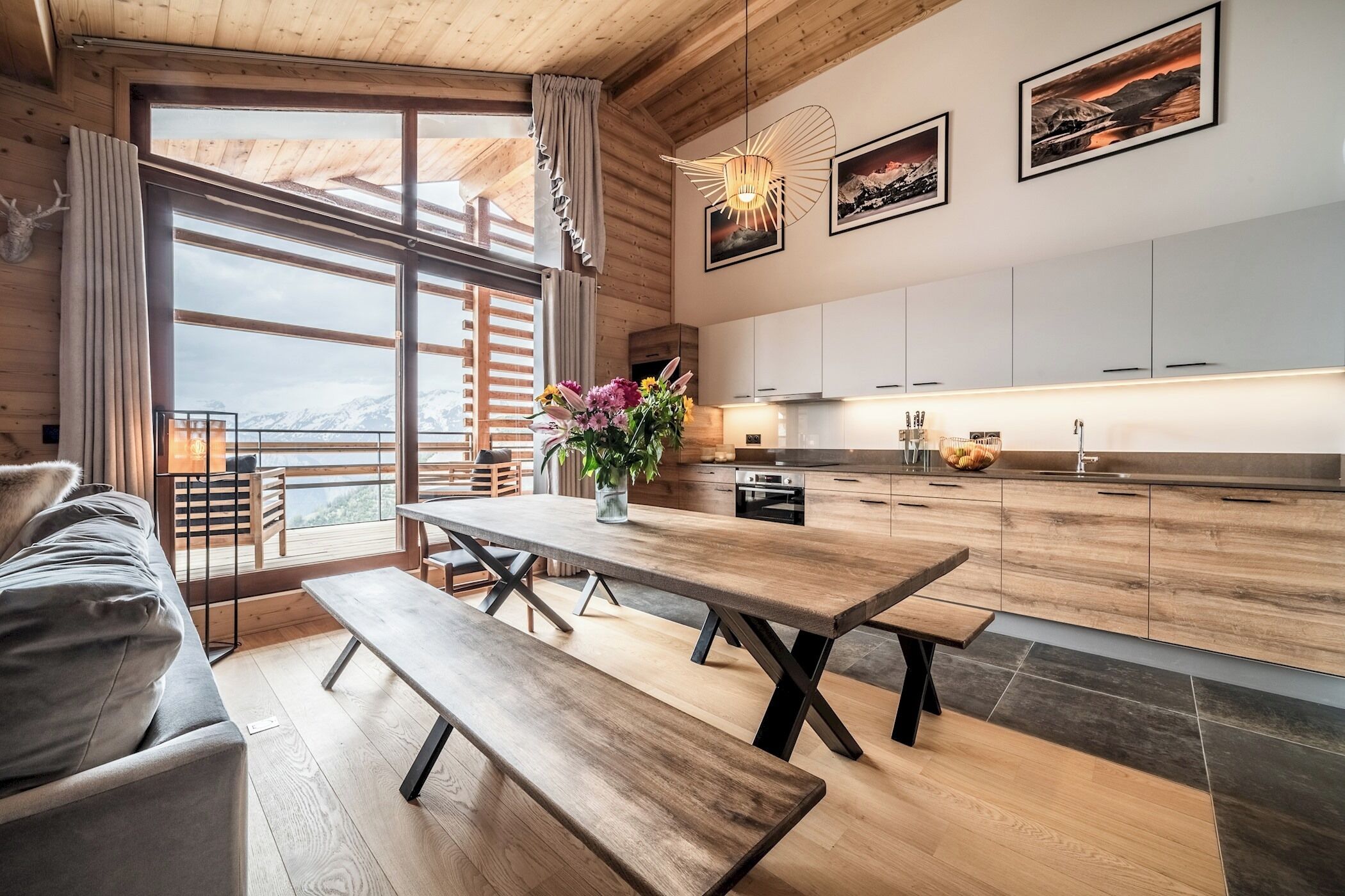 A modern kitchen with wooden cabinets, a dining table with benches, and a large window overlooking a snowy mountain.