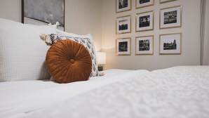 A neatly made bed with white bedding and a brown accent pillow, a bedside table with a lamp, and a wall decorated with framed black-and-white photographs. - Cottage 2 mi to Broadway: King Bed, Fenced Yard (Nashville)