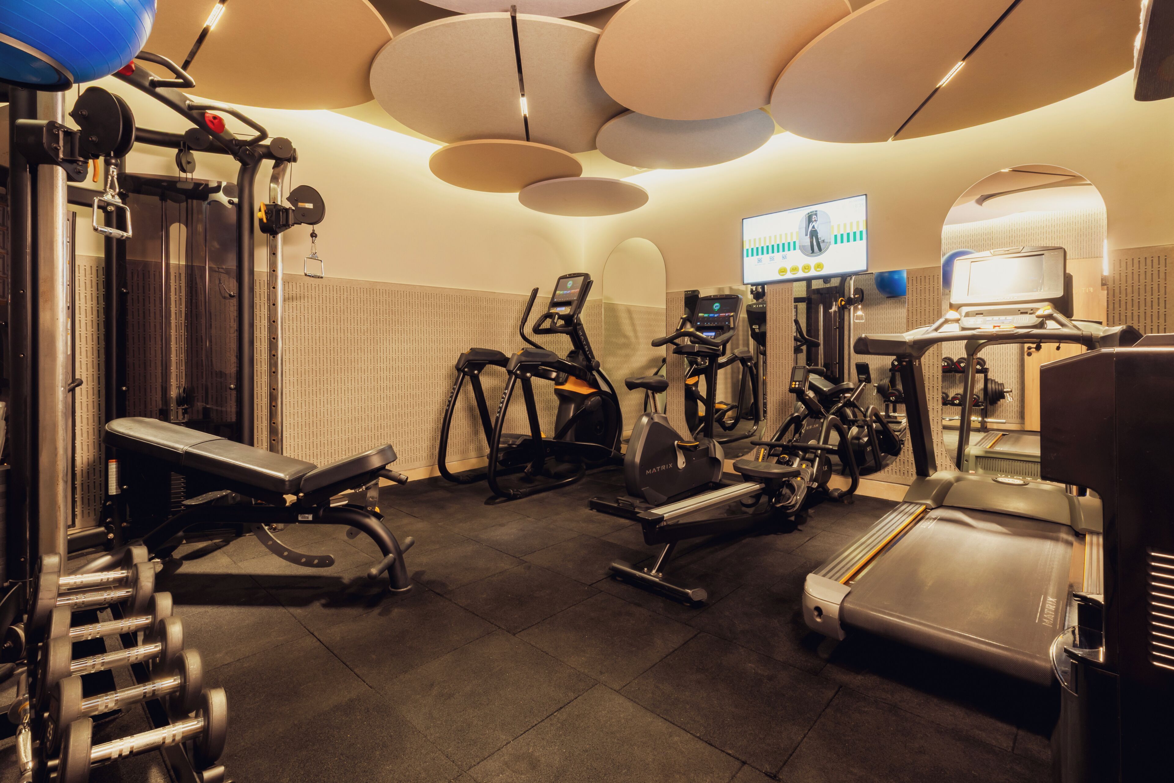 A gym with exercise equipment, a treadmill, and a wall-mounted screen displaying workout metrics.