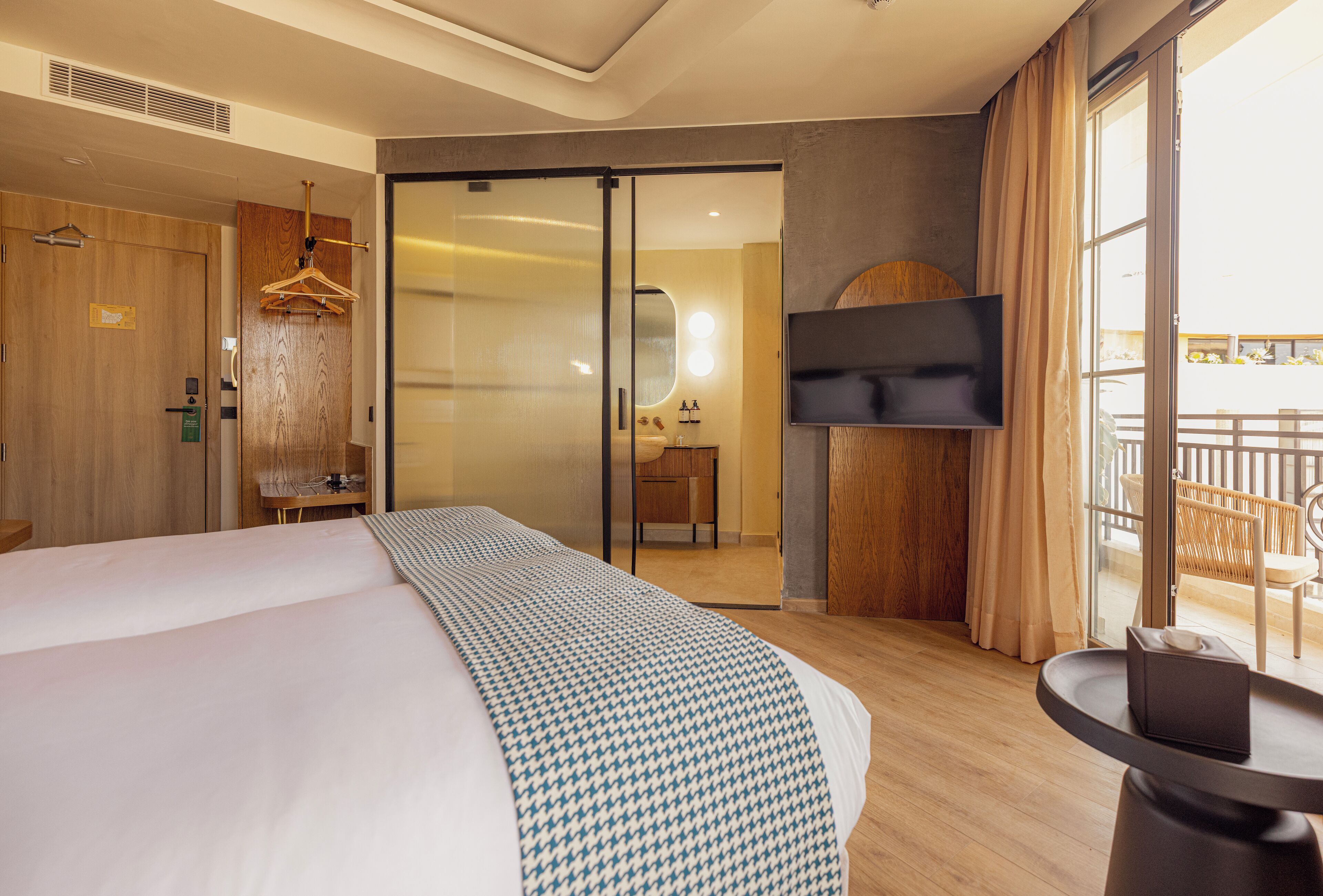 Habitación de hotel con una cama grande, un escritorio con silla, un televisor montado en la pared y un balcón con mobiliario exterior.