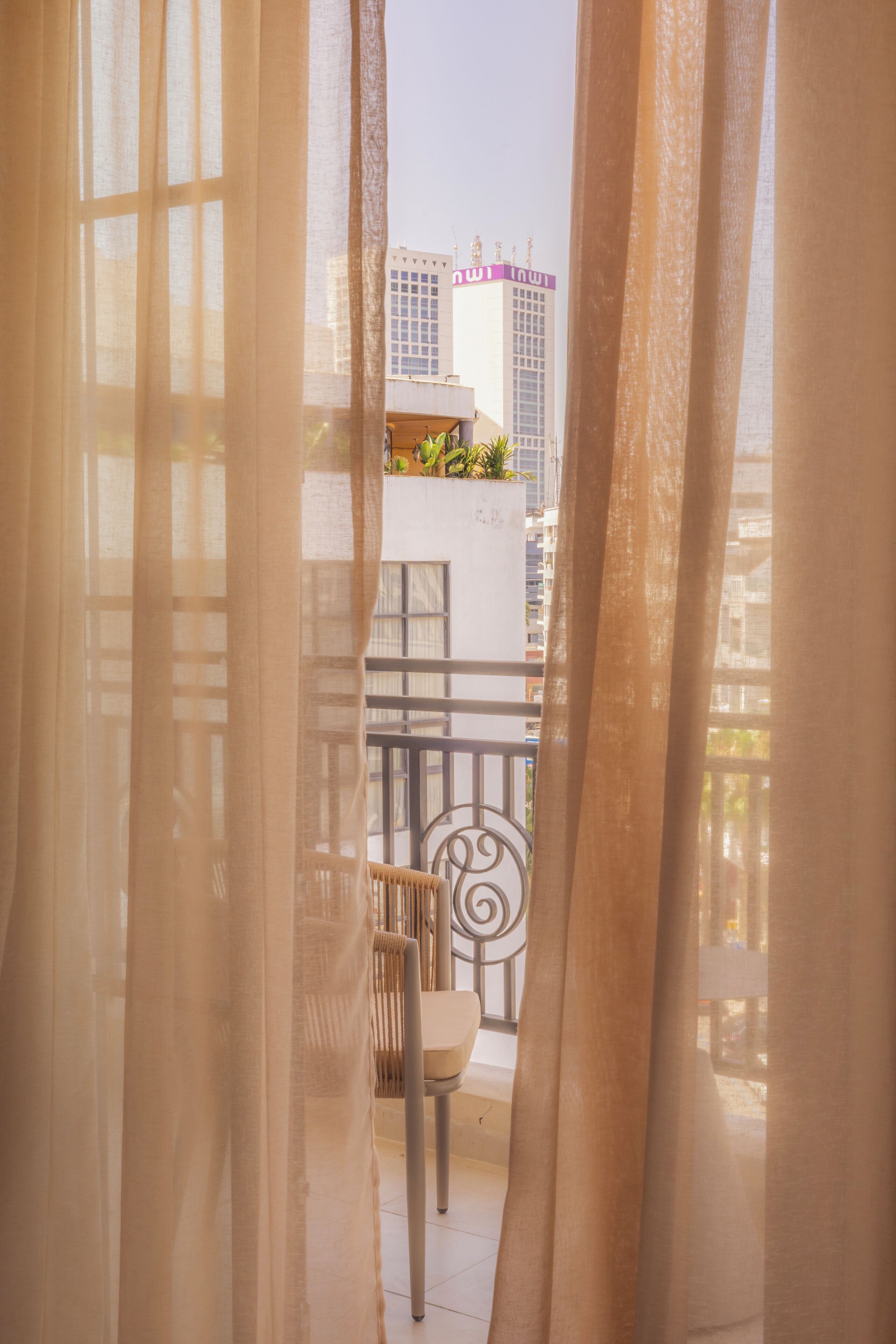 A balcony with a chair and a table, curtains partially drawn, and a view of buildings in the background.