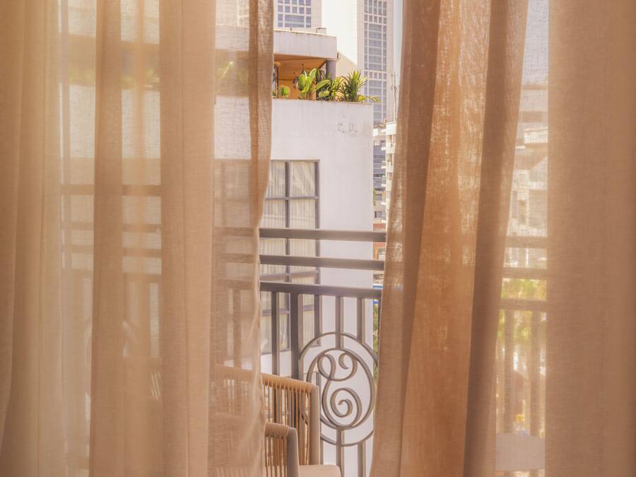 A balcony with a chair and a table, curtains partially drawn, and a view of buildings in the background.