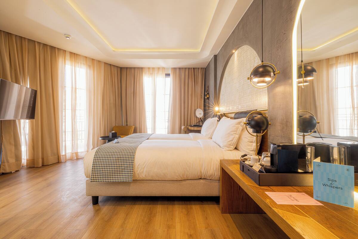 Une chambre d’hôtel avec un grand lit, un bureau équipé d’un ordinateur et d’un téléphone, une télévision, et un panneau indiquant les équipements disponibles dans la chambre.