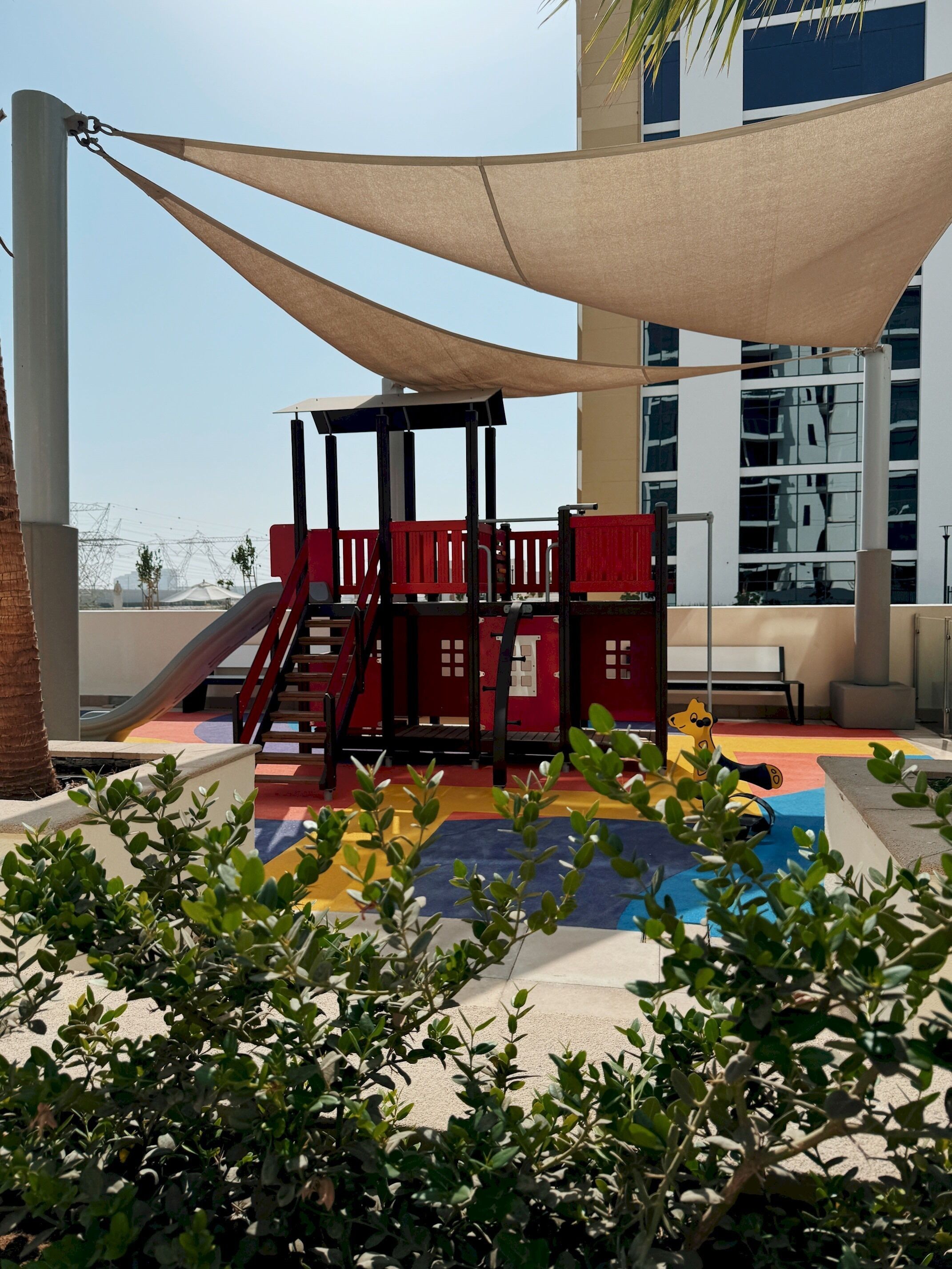 Ein Spielplatz mit Rutsche und Klettergerüsten, umgeben von Grünflächen und einem Gebäude im Hintergrund.