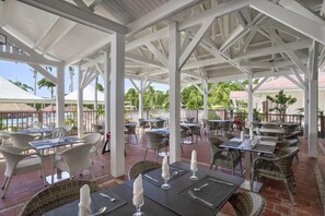 Uma área de estar ao ar livre com mesas e cadeiras, piscina ao fundo e uma estrutura de gazebo branca.