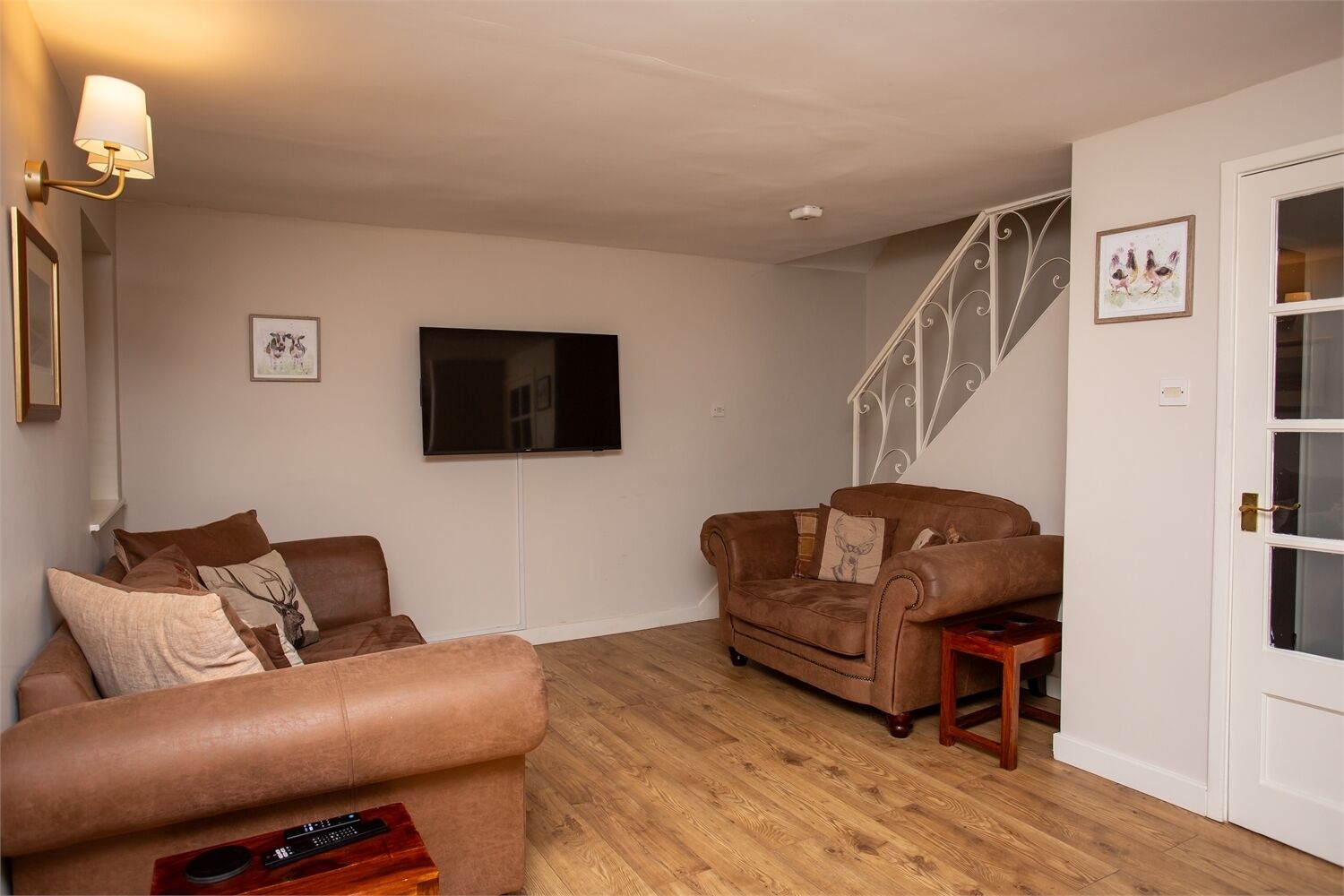 A living room with two brown sofas, a small wooden table, a TV mounted on the wall, and a staircase with a decorative railing.