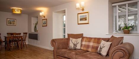 A living room with a brown sofa, wooden dining table, and chairs. There are wall-mounted lights and framed pictures.