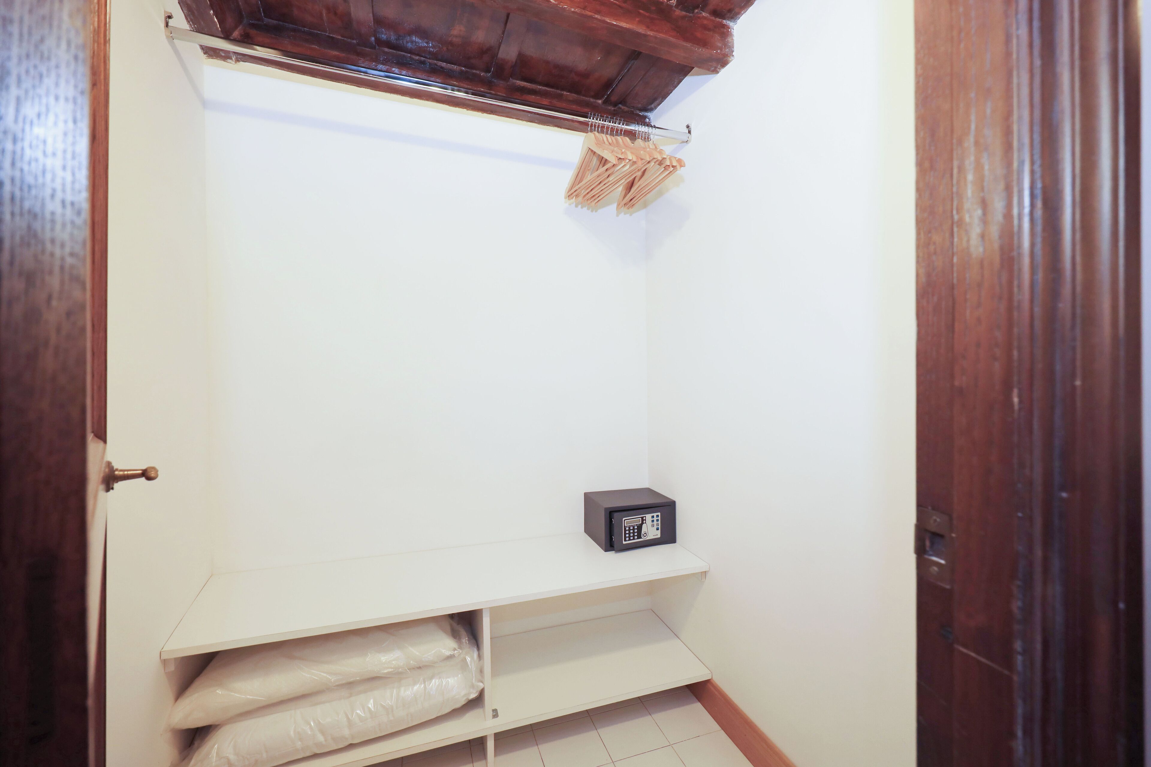 A small, narrow hallway with a wooden door on the right, a white shelf with bags, and a small black object on the shelf.