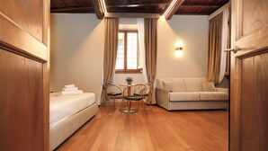 A room with a wooden floor, a bed, a sofa, a small table with chairs, and a window with curtains. - Golden Pantheon Rooms (Rome)