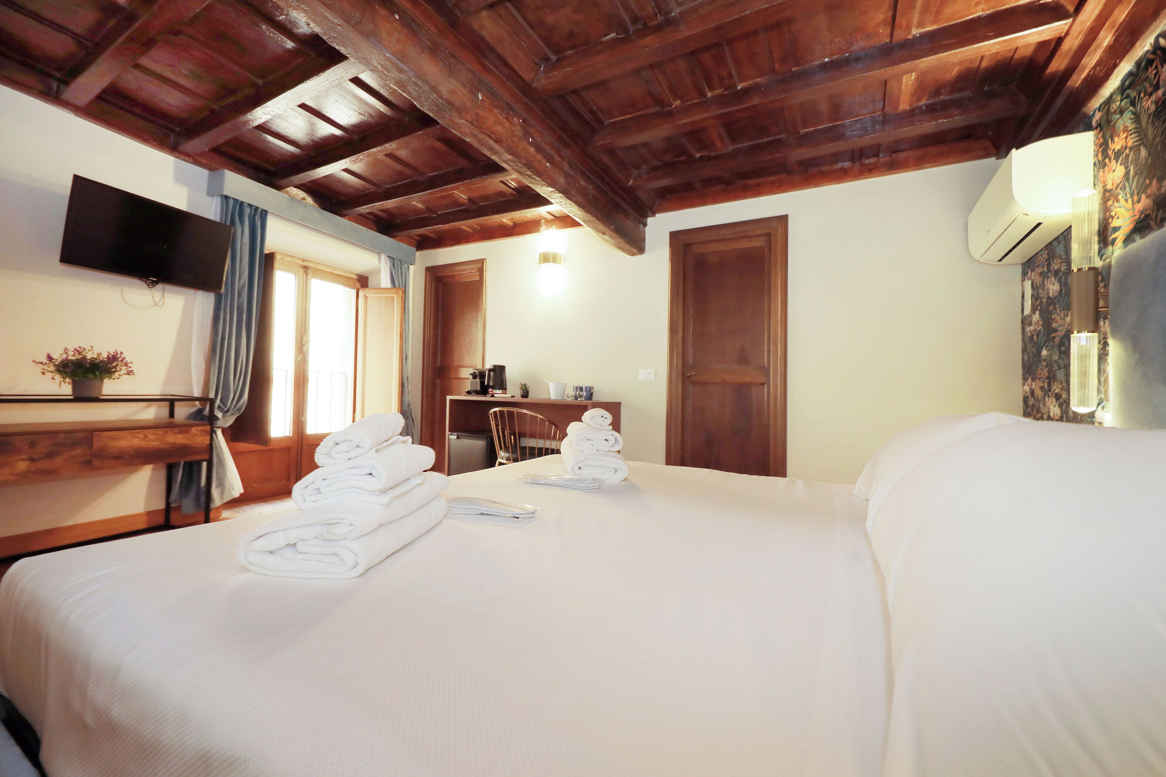 A hotel room with a wooden ceiling, a bed with white linens, a desk with a chair, a flat-screen TV, and an air conditioning unit.