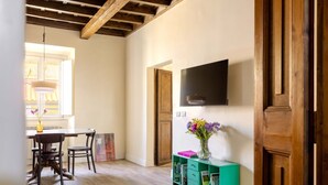 Ein Zimmer mit Esstisch, Stühlen, einem an der Wand montierten Fernseher, einer Holzkommode und einem Fenster mit Vorhängen.