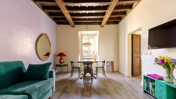 Ein Zimmer mit einem grünen Sofa, einem Esstisch mit Stühlen, einem Fernseher an der Wand und einer Holzbodenverkleidung.