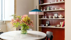 Une table ronde ornée d’un vase de fleurs, un parasol bleu et rose, et une bibliothèque contenant des objets décoratifs.