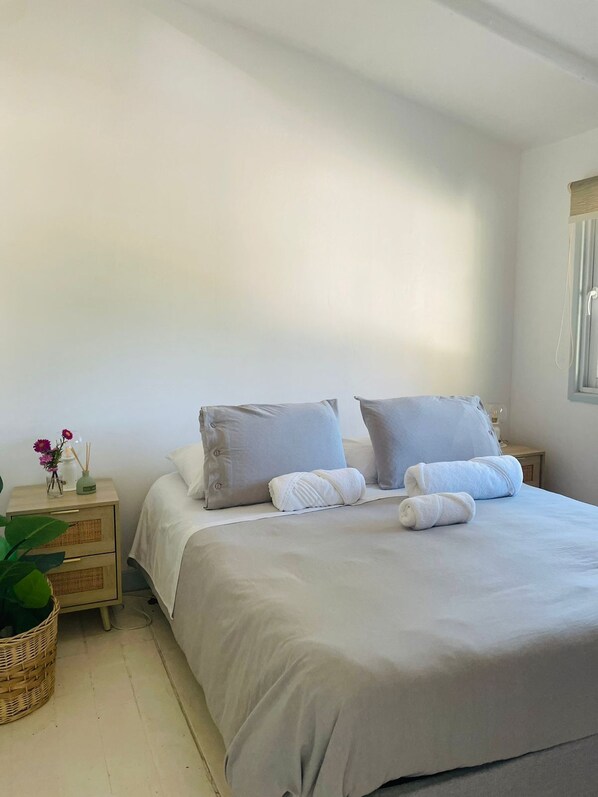 A neatly made bed with white linens and two pillows, a bedside table with a vase of flowers, and a wicker basket on the floor.